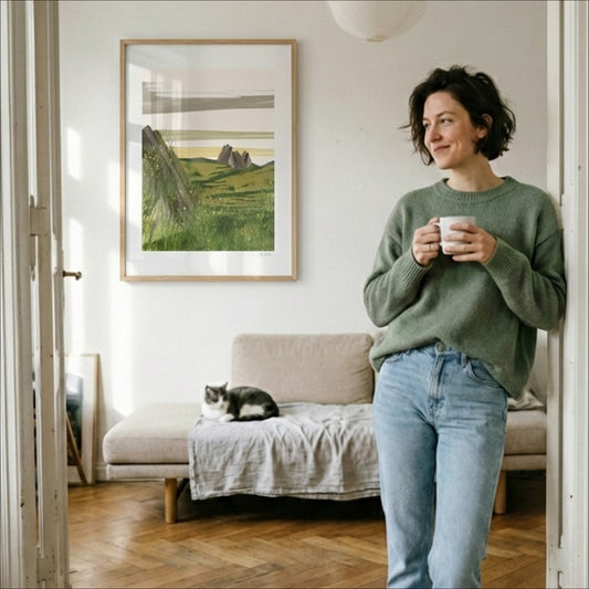Open Field art print leaning against a sofa cushion in a cozy Scandinavian interior, OAM Studio and Woman in a green sweater holding a mug in a living room with a cat on the couch and a framed picture on the wall.